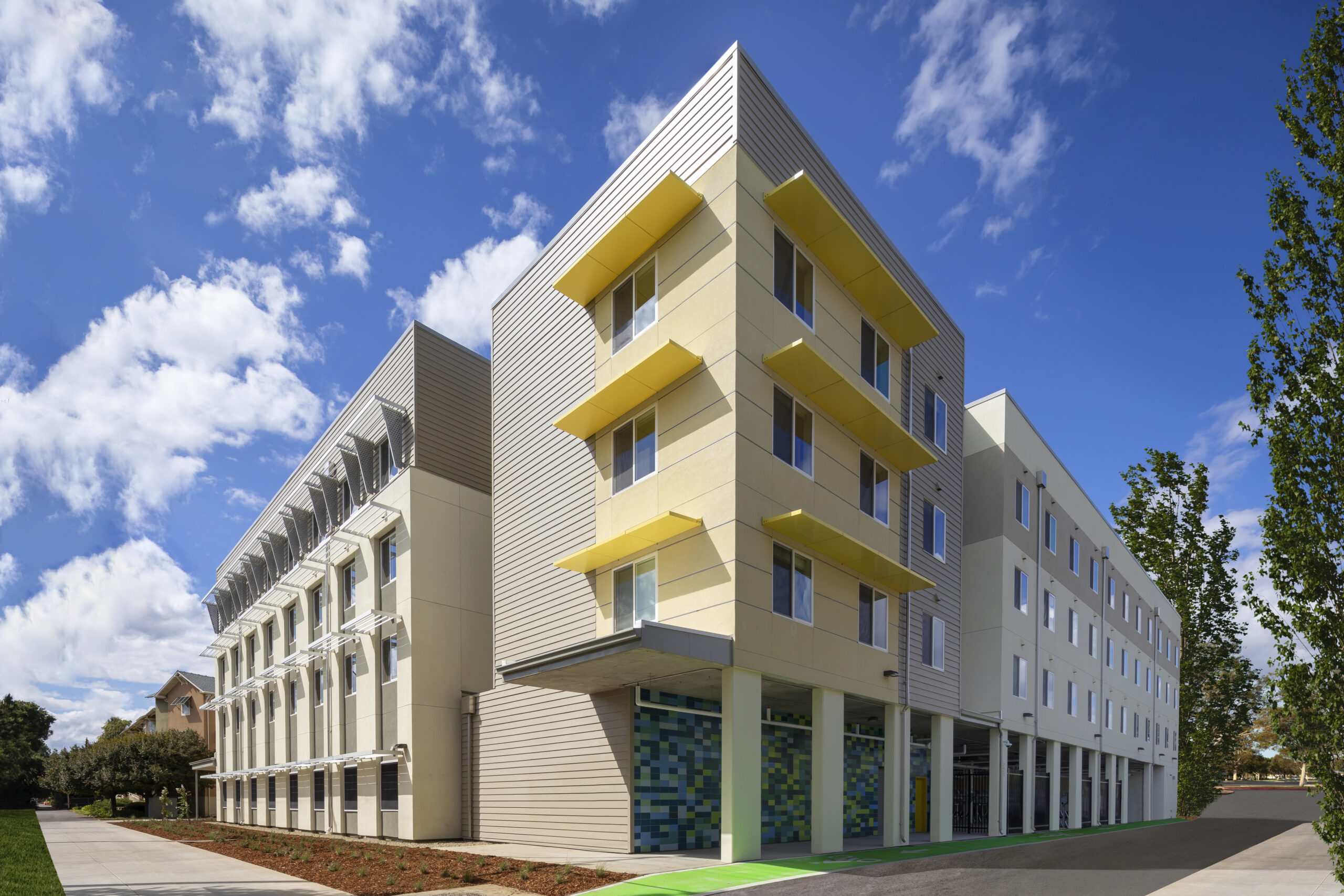Modern multi-story building with yellow awnings, large windows, and a landscaped walkway under a blue sky.