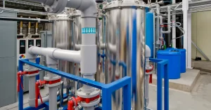 Close-up view of a modern HVAC and plumbing system in a healthcare or industrial facility, featuring stainless steel tanks, PVC piping, red valve controls, and blue chemical storage barrels.